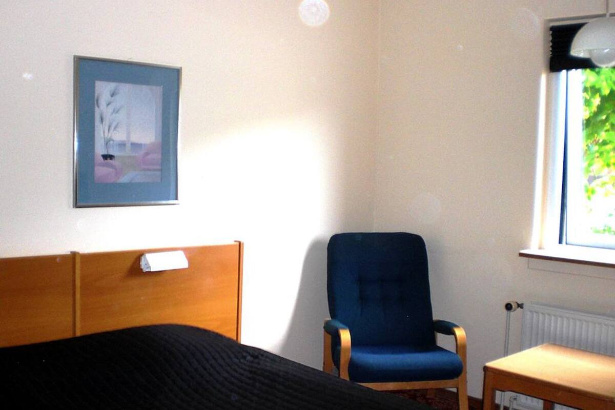 A room with a bed, a blue chair, a small table, a framed picture on the wall, and a window with a view of greenery.