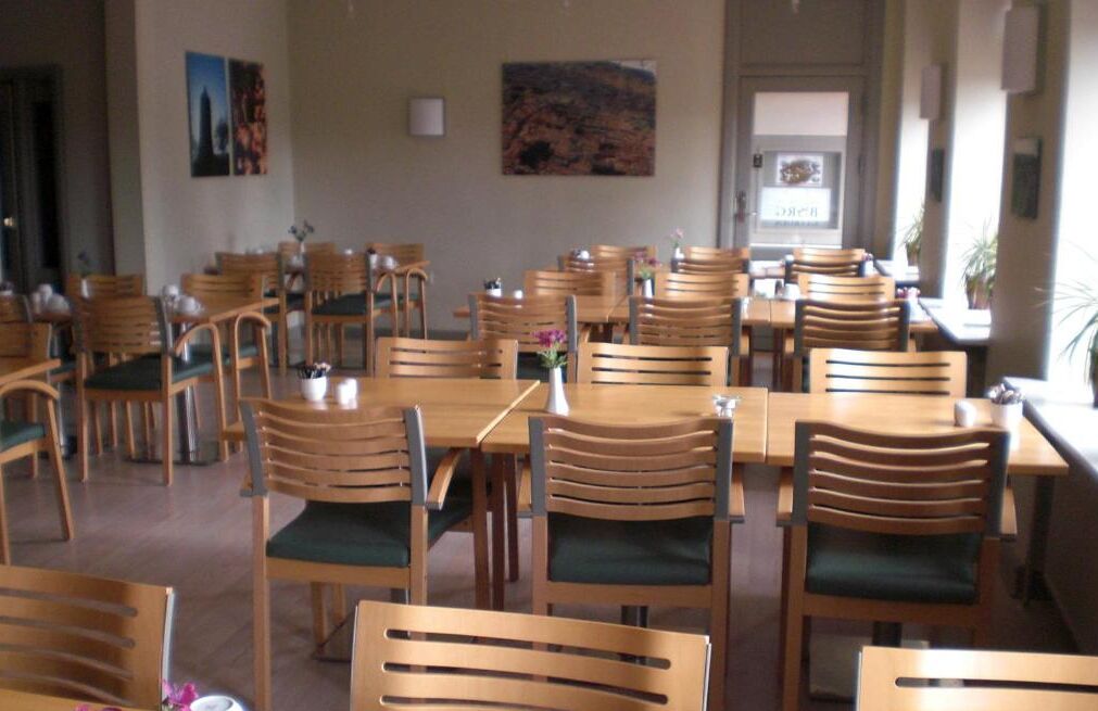 A dining area with wooden tables and chairs, some with green cushions. There are framed pictures on the walls.