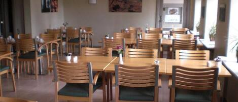 A dining area with wooden tables and chairs, some with green cushions. There are framed pictures on the walls.
