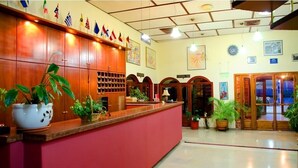 A reception area with a red counter, wooden cabinets, and multiple potted plants. Flags are displayed on the wall above the counter. - Oscar Hotel (Igoumenitsa)