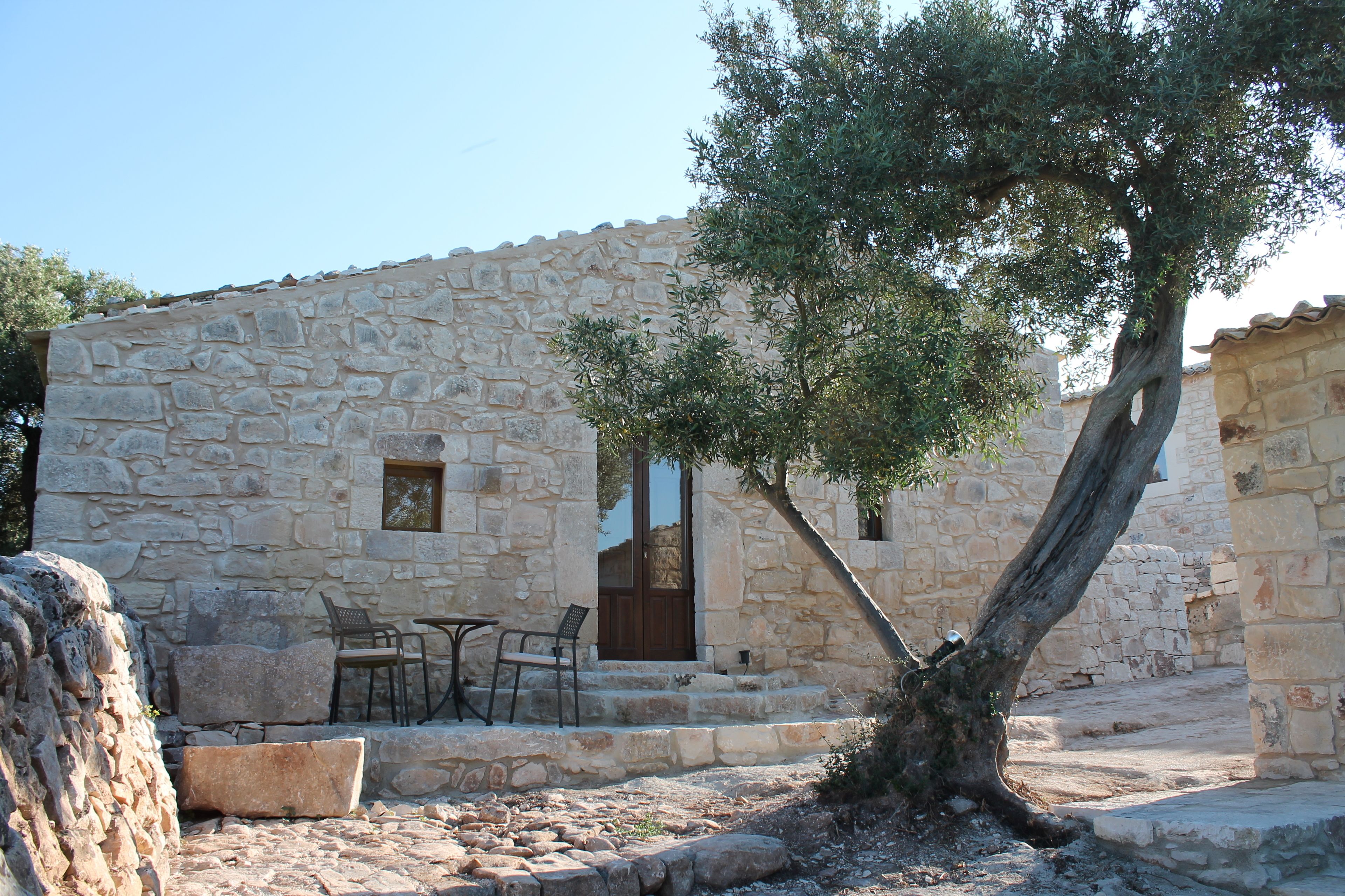 A stone building with a wooden door, a small window, and a tree in front. There is a table and two chairs outside.