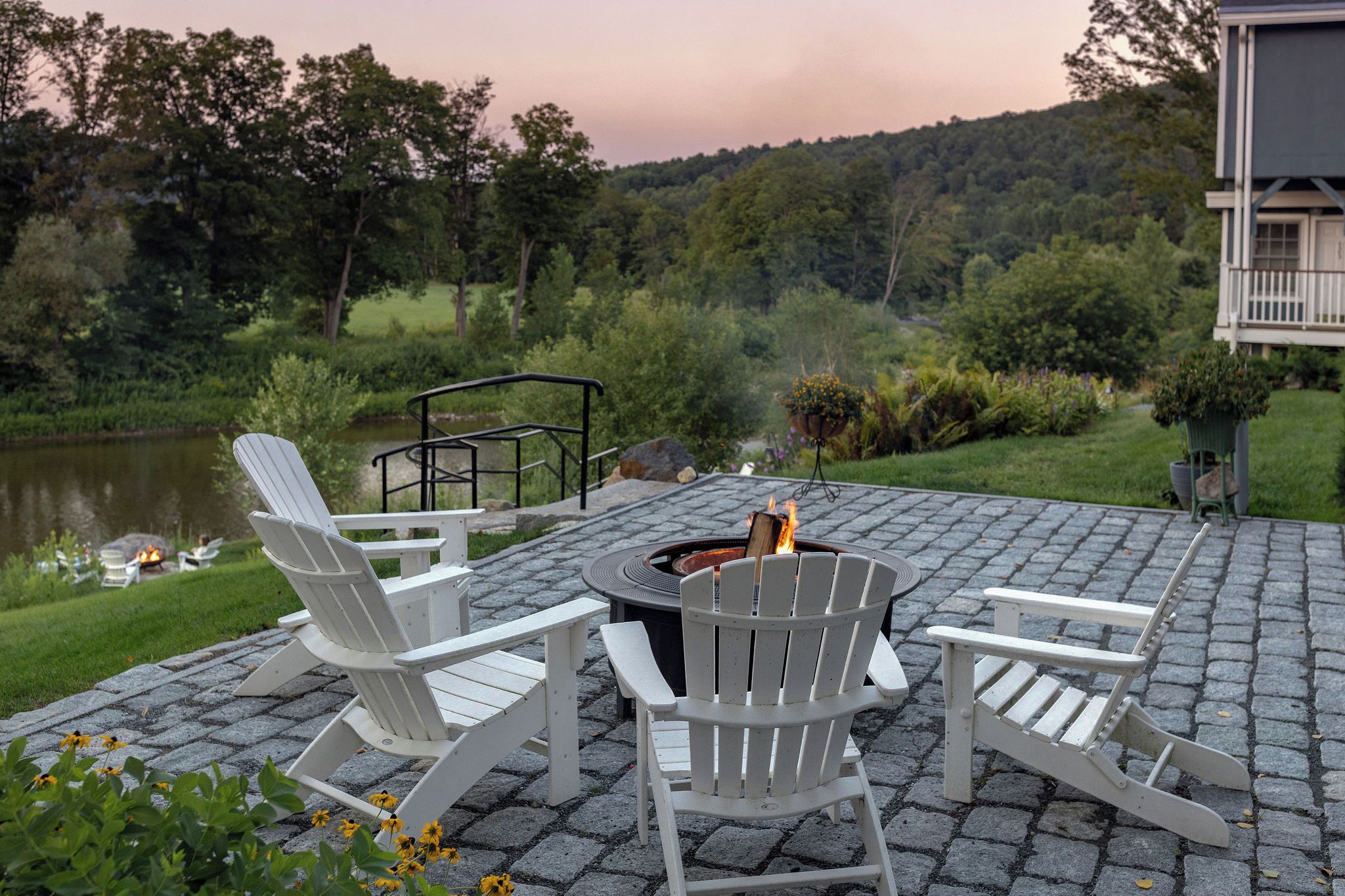 An outdoor seating area with Adirondack chairs, a fire pit, and a view of a lake and forest.