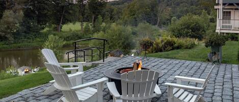 An outdoor seating area with Adirondack chairs, a fire pit, and a view of a lake and forest.