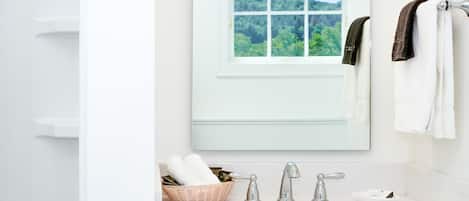 Une salle de bain avec un lavabo blanc, deux robinets, un miroir et une fenêtre donnant sur un paysage verdoyant.