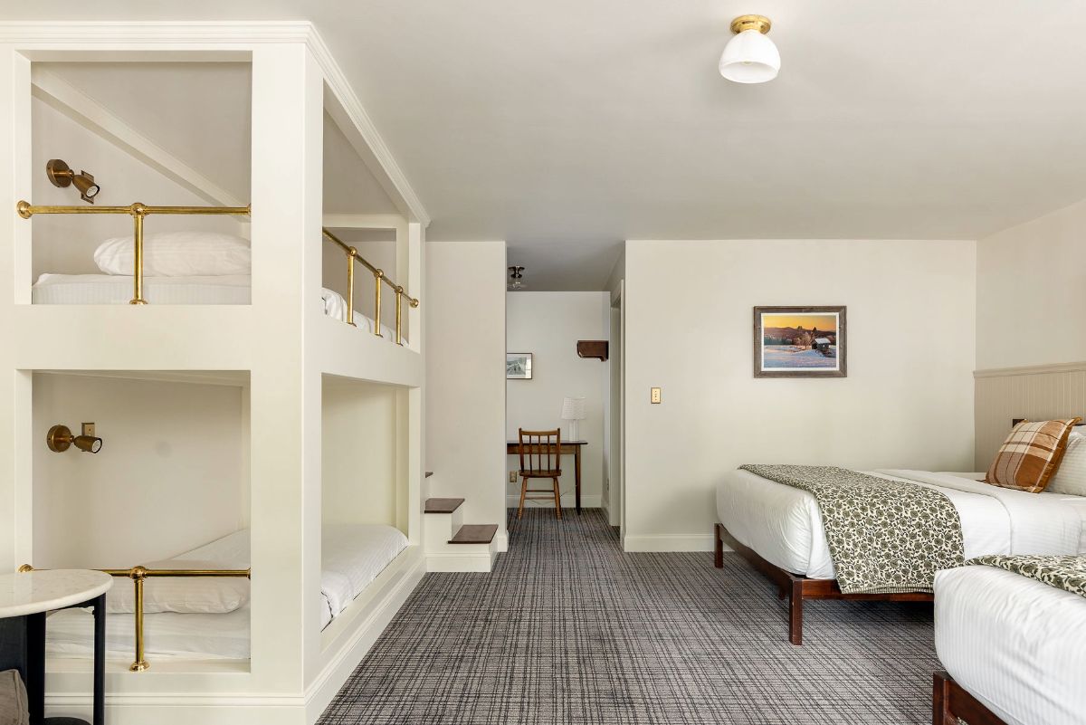 A hotel room with a bunk bed, a desk, a chair, and a bed with a patterned throw.