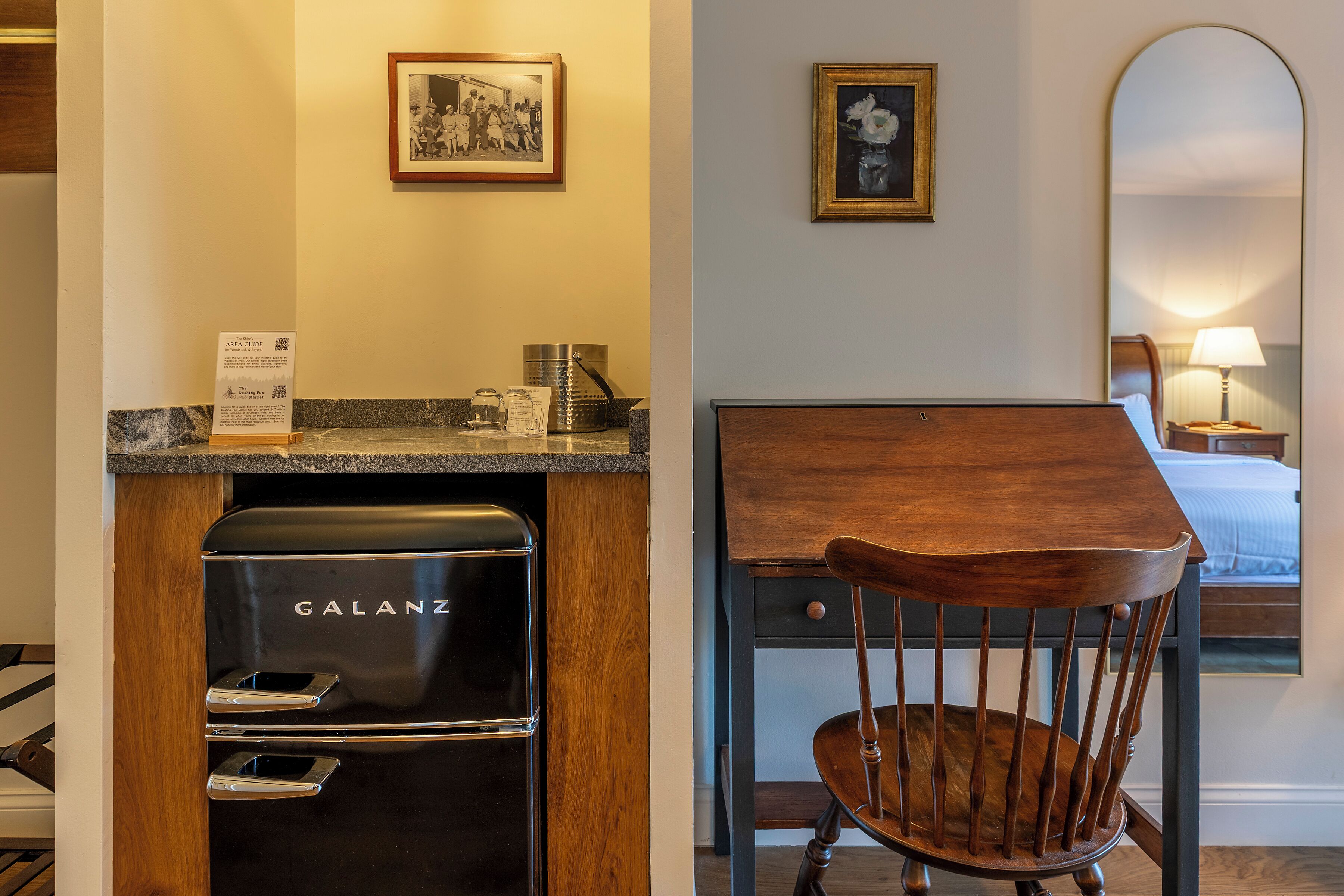 A hotel room with a black Galanz refrigerator, a wooden chair, a small table, a bed, and a wall-mounted mirror.
