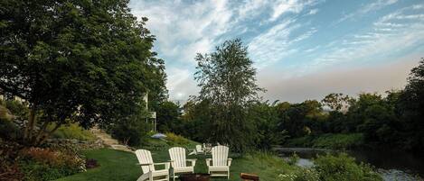 A serene outdoor seating area with Adirondack chairs and a fire pit, surrounded by lush greenery and a calm body of water.