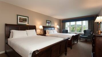 A hotel room with two beds, a desk, a chair, and a window with a view of greenery.
