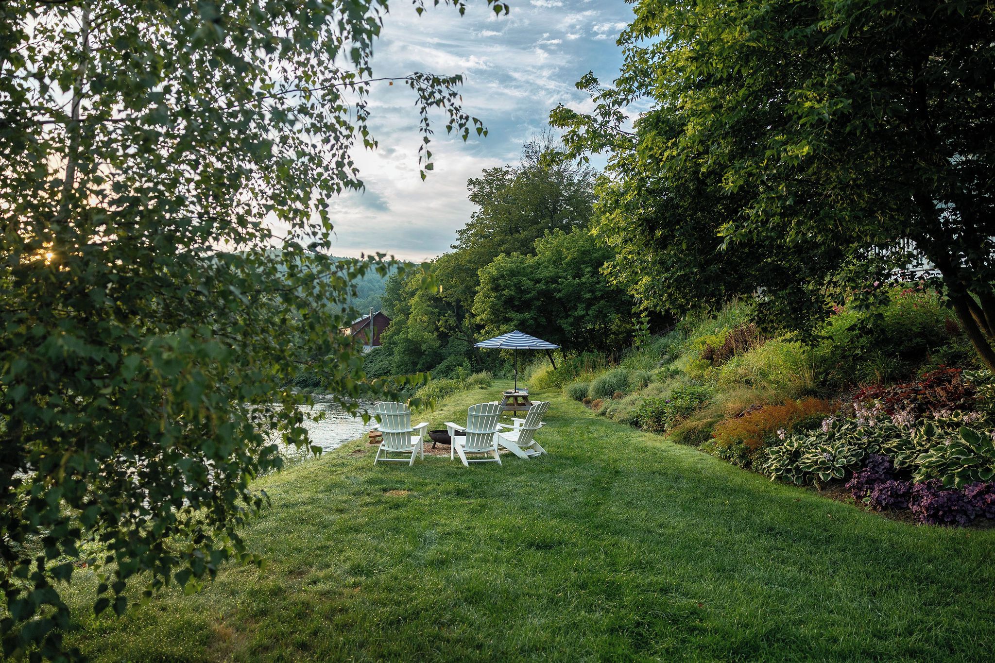 A grassy outdoor area with a pond, surrounded by trees and shrubs. There are white Adirondack chairs and a blue umbrella.