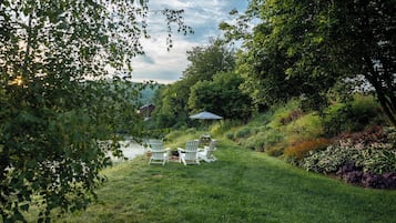 A grassy outdoor area with a pond, surrounded by trees and shrubs. There are white Adirondack chairs and a blue umbrella.