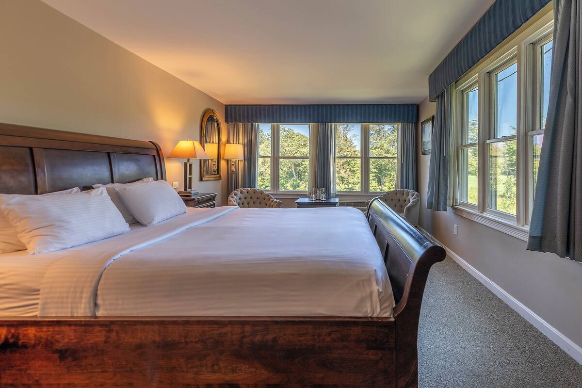 Una cama bien hecha con cabecera de madera, una mesita de noche con lámpara y vistas a árboles a través de grandes ventanales.