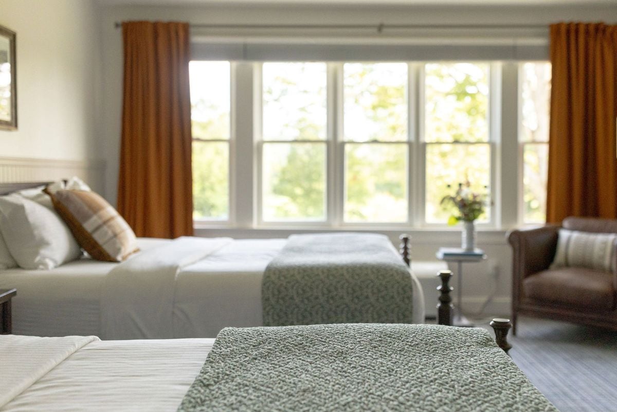 Quarto com duas camas, janela com cortinas, uma mesinha com um vaso e uma cadeira.