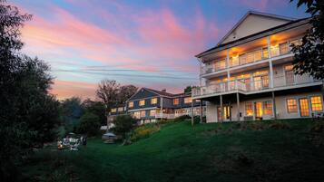 A multi-story building with a balcony, surrounded by greenery and a sunset sky.