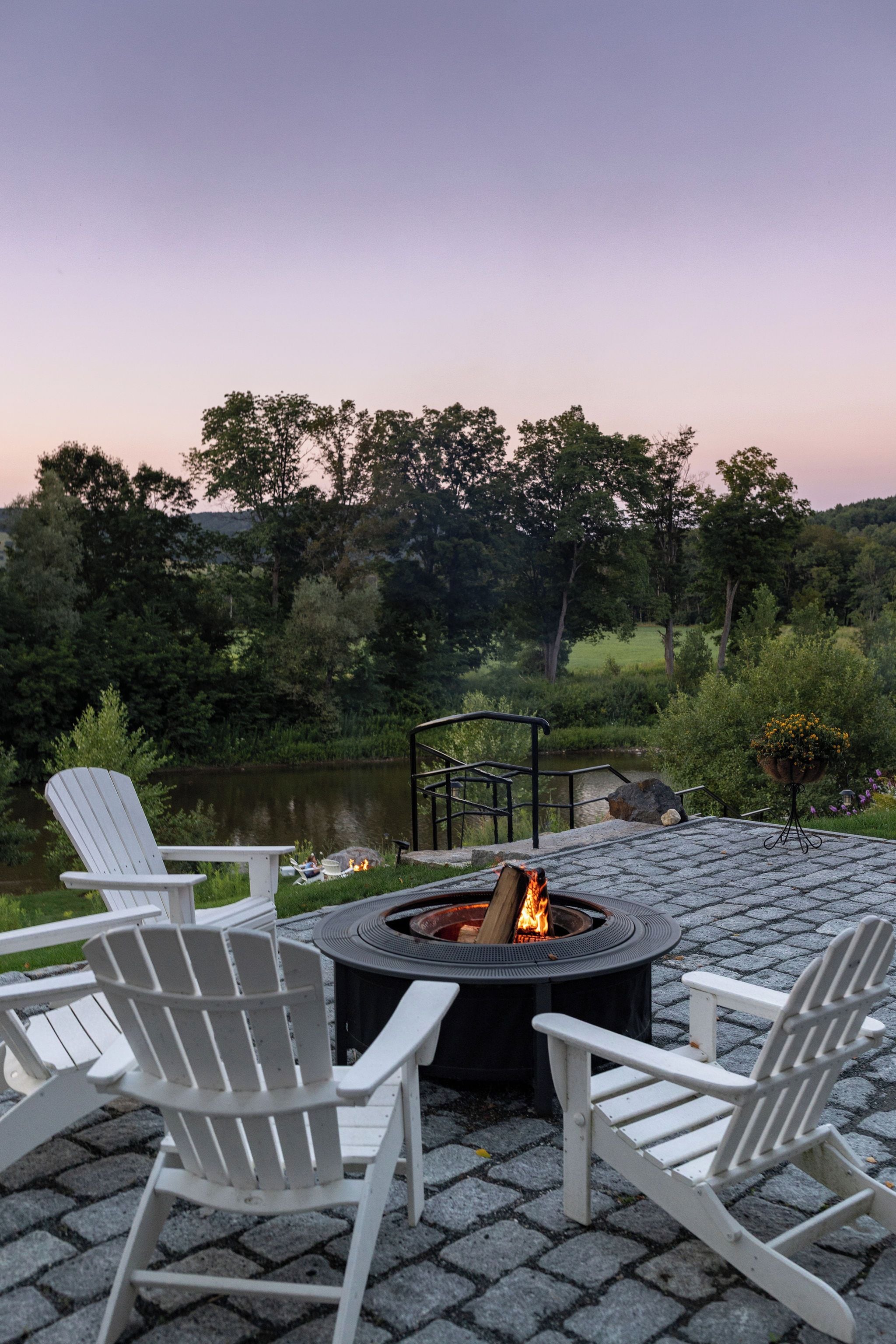 A fire pit surrounded by Adirondack chairs on a paved patio with a view of a lake and trees.