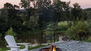 A fire pit surrounded by Adirondack chairs on a paved patio with a view of a lake and trees.