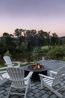 Un pozo de fuego rodeado de sillas Adirondack en un patio empedrado con vista a un lago y árboles.