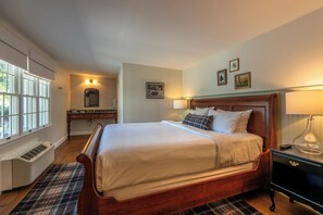 Un dormitorio ordenado con una cama grande, una mesita de noche con lámpara, un aparador y una ventana con persianas.