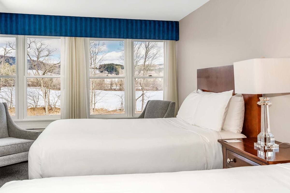 Habitación de hotel con una cama grande, una mesita de noche con lámpara, una silla y vistas a un paisaje nevado a través de grandes ventanales.