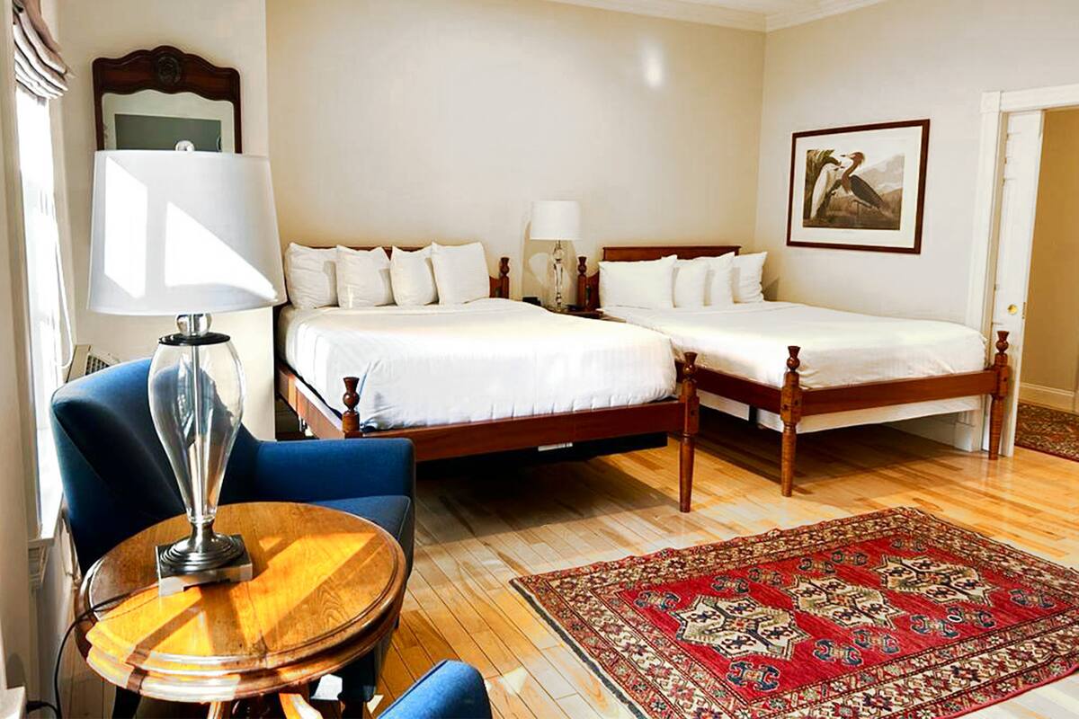 Una habitación de hotel con dos camas individuales, una silla de madera, una mesa redonda de madera, un tapete rojo estampado y un cuadro en la pared.