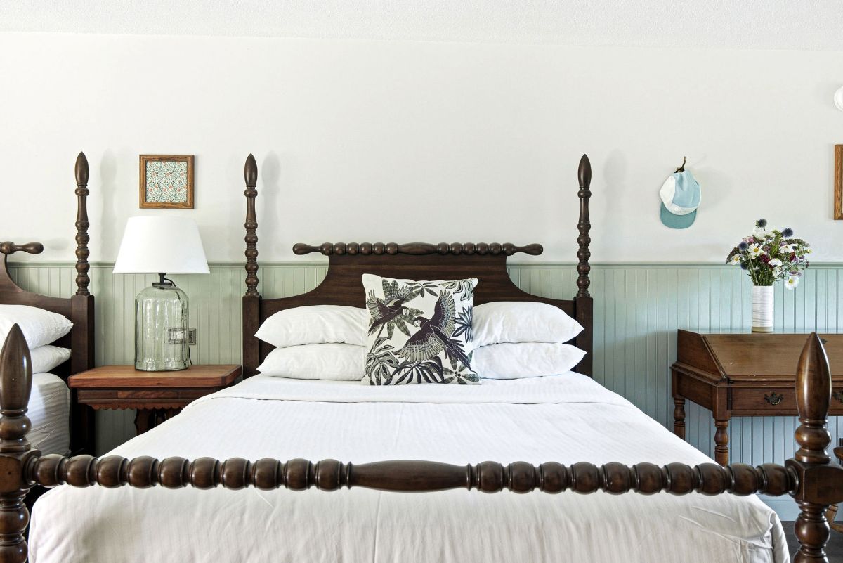Una cama con dosel, cabecero decorativo, ropa de cama blanca, mesita de noche con lámpara y perchero de pared.