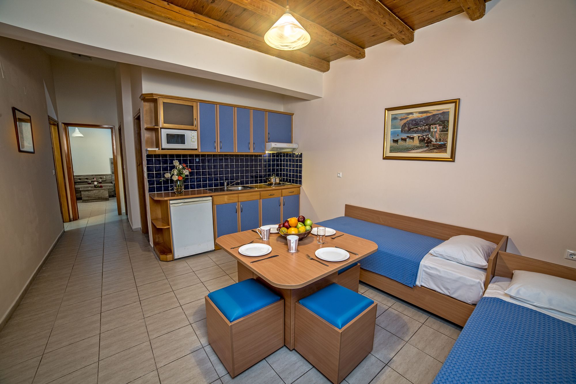 A compact room with a kitchenette, a dining table with blue cushioned chairs, a bed with blue bedding, and a painting on the wall.