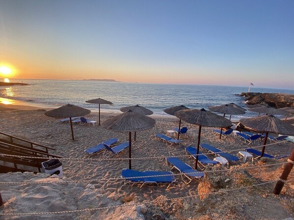 A beach with sun loungers and umbrellas, a staircase leading down to the beach, and a flag on a rock.