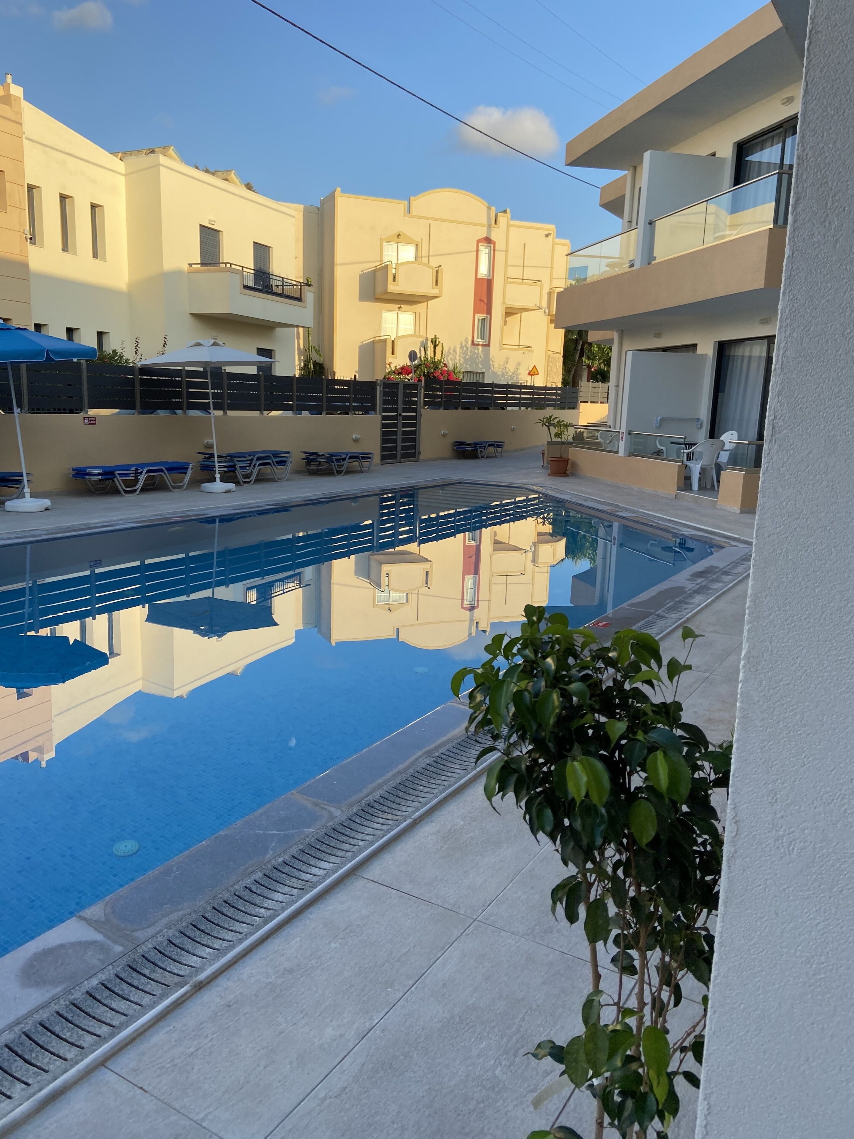 En swimmingpool med solstolar och parasoller, omgiven av byggnader med balkonger och fönster.