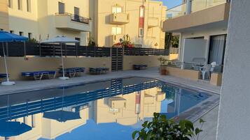 En swimmingpool med solstolar och parasoller, omgiven av byggnader med balkonger och fönster.