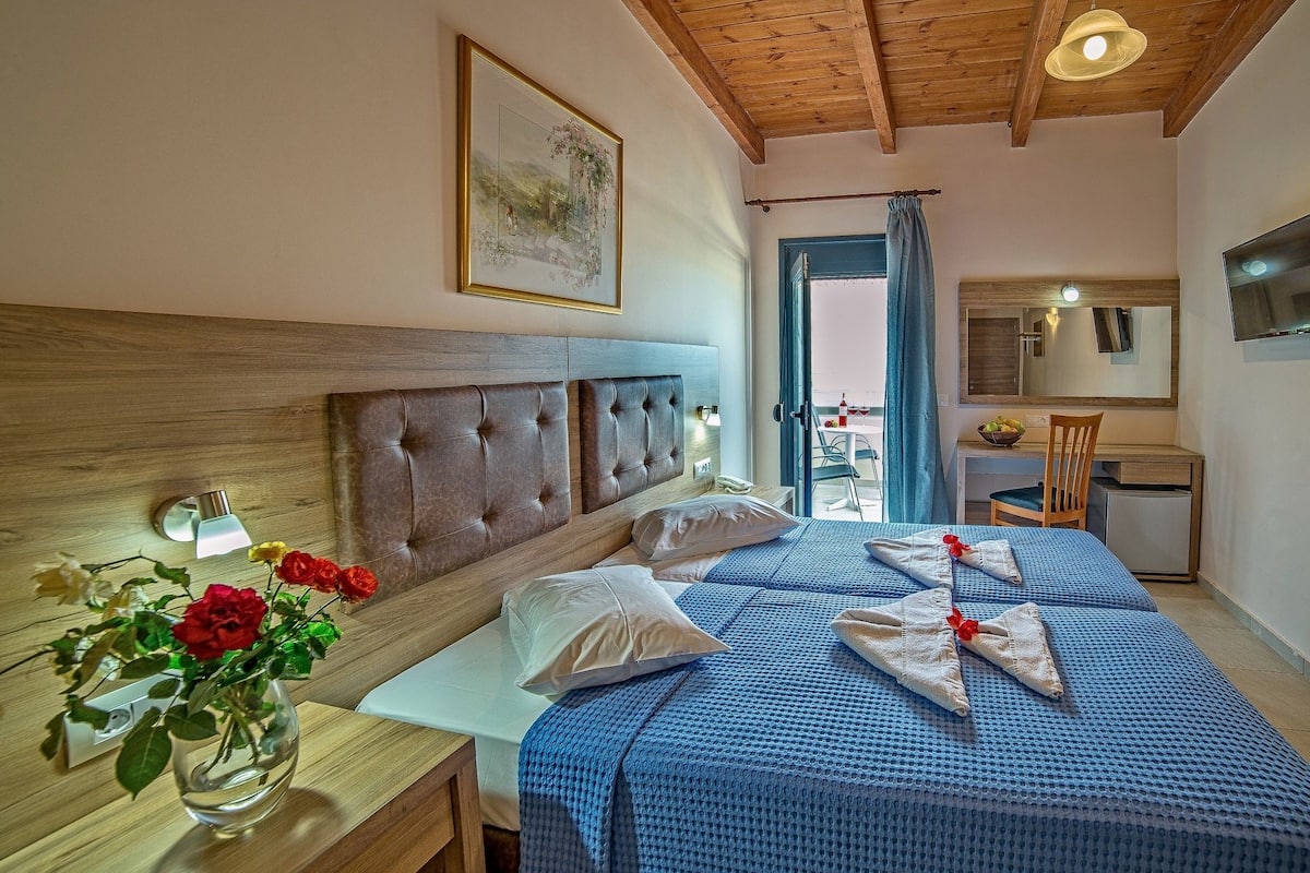 Ein Schlafzimmer mit einem Holz-Kopfteil, einem Nachttisch mit Blumen in einer Vase, einem Bett mit blauer Bettwäsche, einem Schreibtisch mit Stuhl und einem Balkon mit Blick.