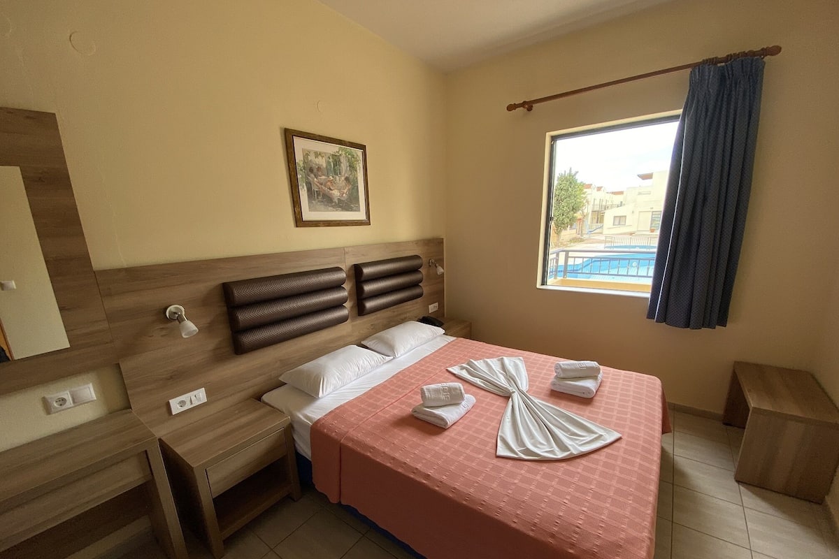 Ein Hotelzimmer mit einem hölzernen Kopfteil, einem Bett mit rosa Bettwäsche, zwei weißen Kissen und einem ordentlich gefalteten Handtuch auf dem Bett.