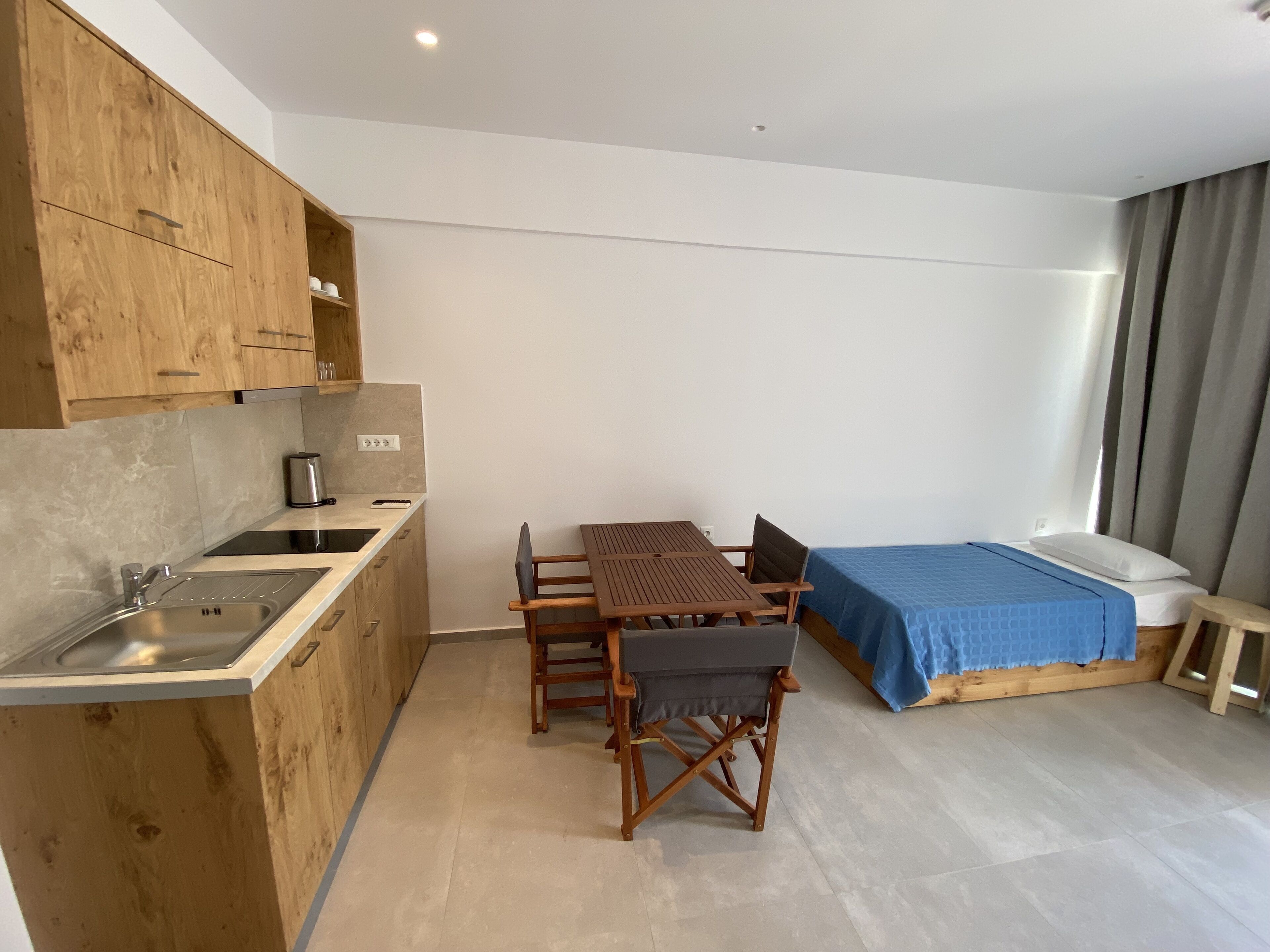 A compact room with a kitchenette, a dining table with chairs, a bed with a blue bedspread, and a small stool.