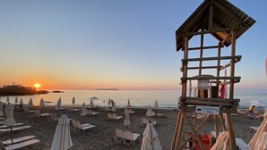 Une plage avec des transats et des parasols, une tour de surveillance pour les sauveteurs, et un coucher de soleil en arrière-plan.