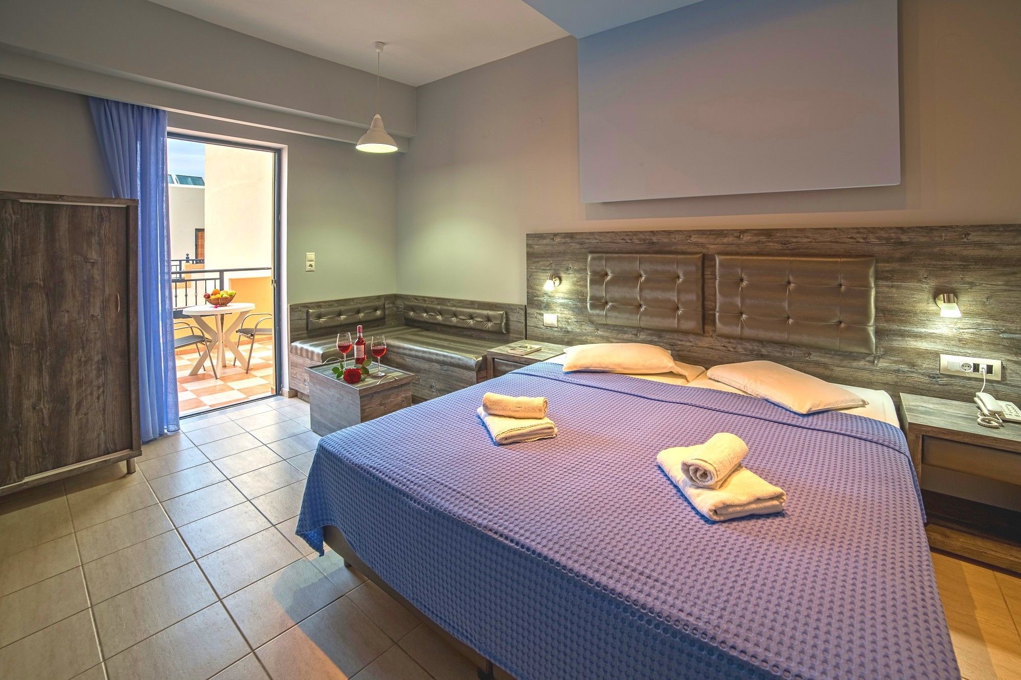 Ein Hotelzimmer mit einem großen Bett, einem Holzkopfende, einem kleinen Tisch mit Flasche und Gläsern, einer Bank und Blick ins Freie.