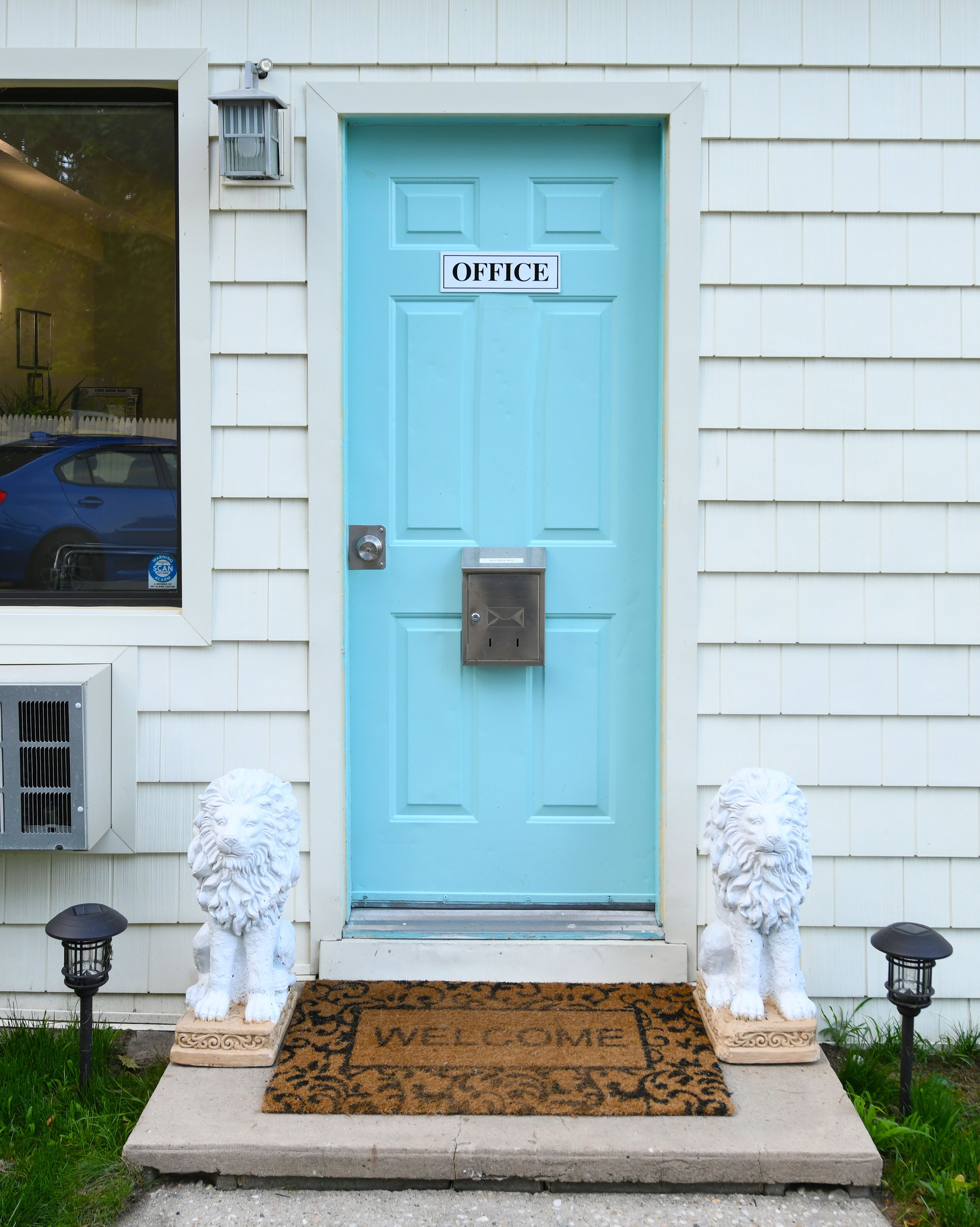 A light blue door with a sign reading "OFFICE" above it, flanked by two white lion statues, and a welcome mat at the entrance.
