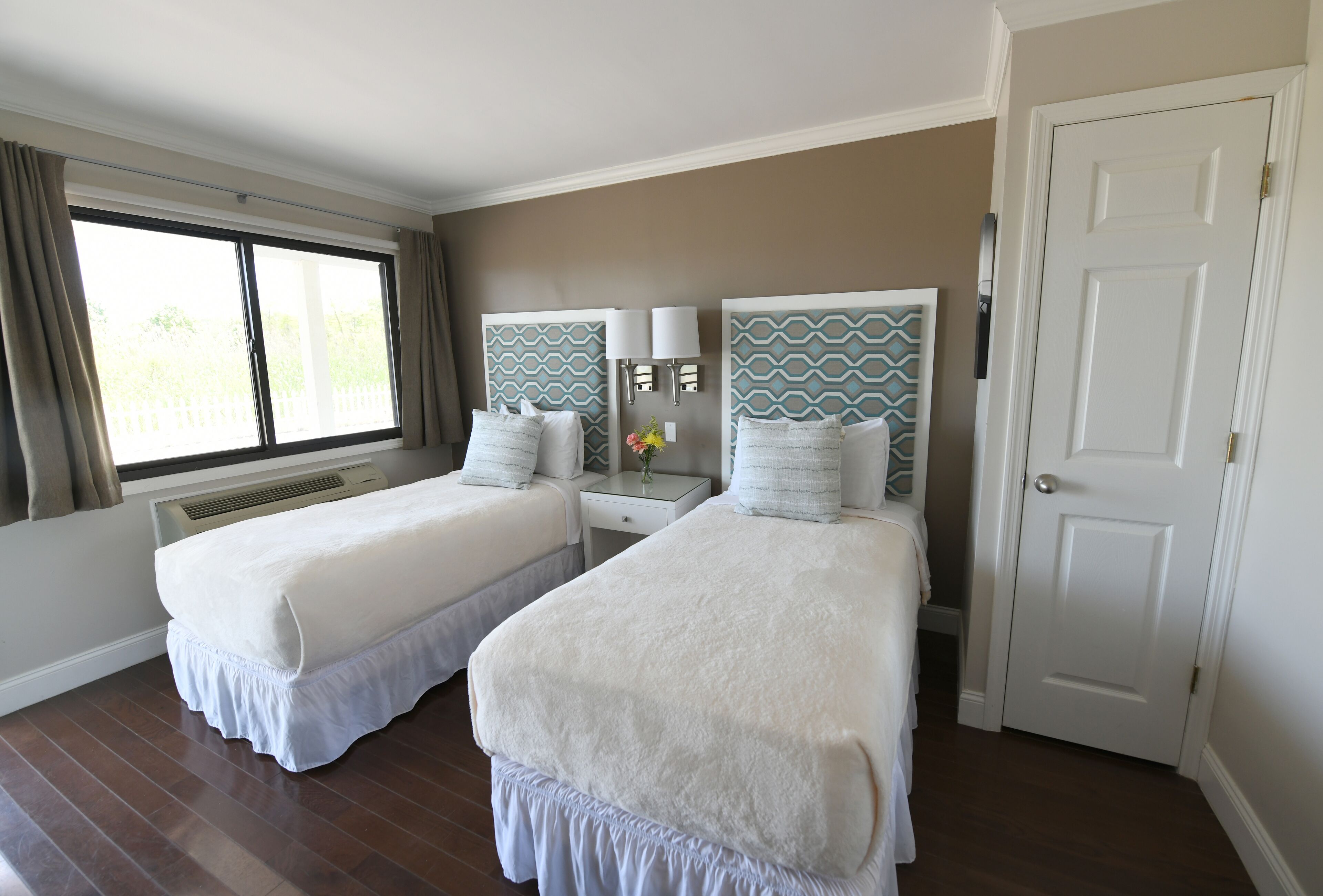 Une chambre d’hôtel avec deux lits, une fenêtre avec des rideaux, une porte blanche et une lampe fixée au mur.