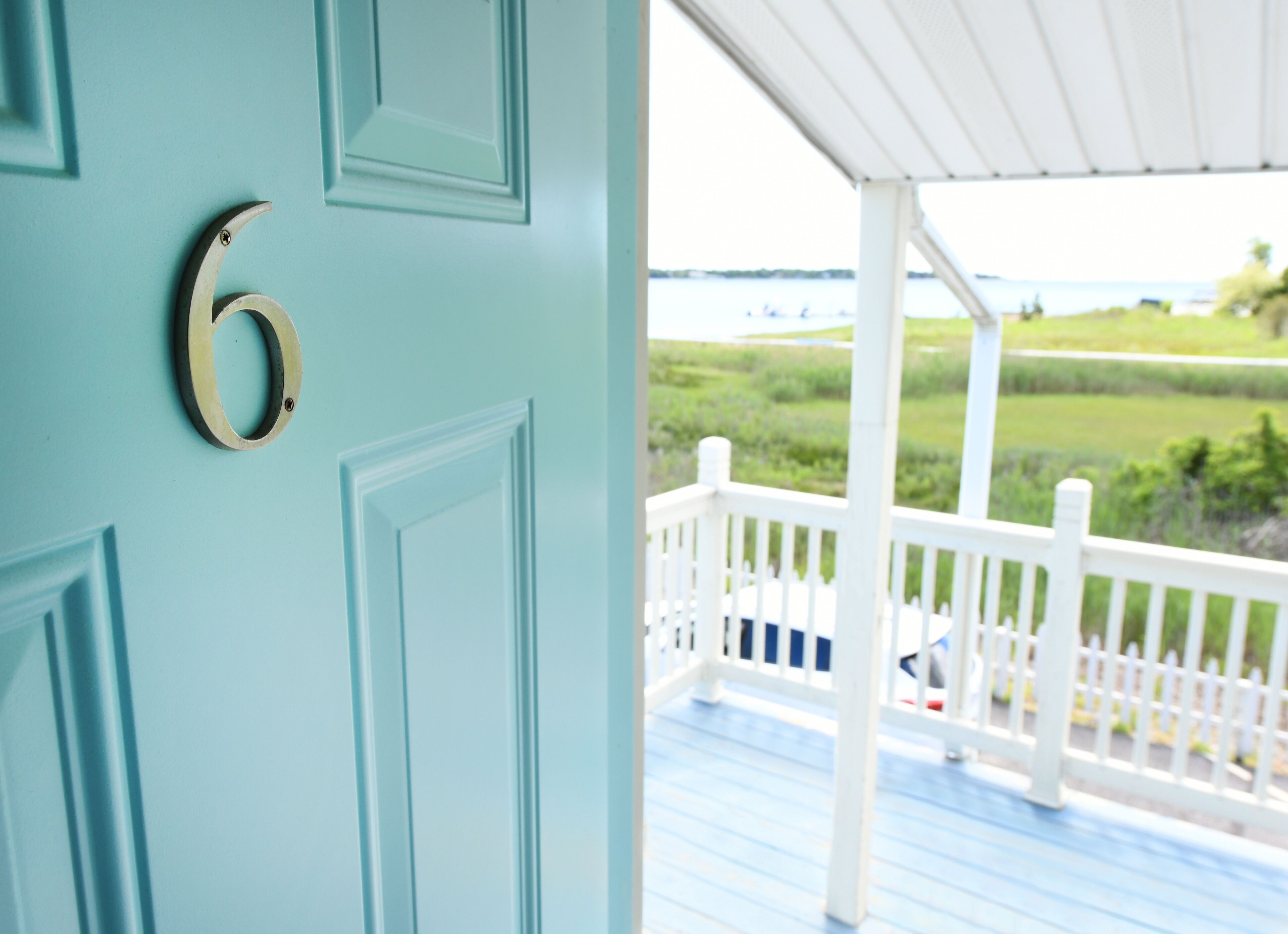 A teal door with a brass number six, leading to a porch with a white railing and a view of a grassy landscape.