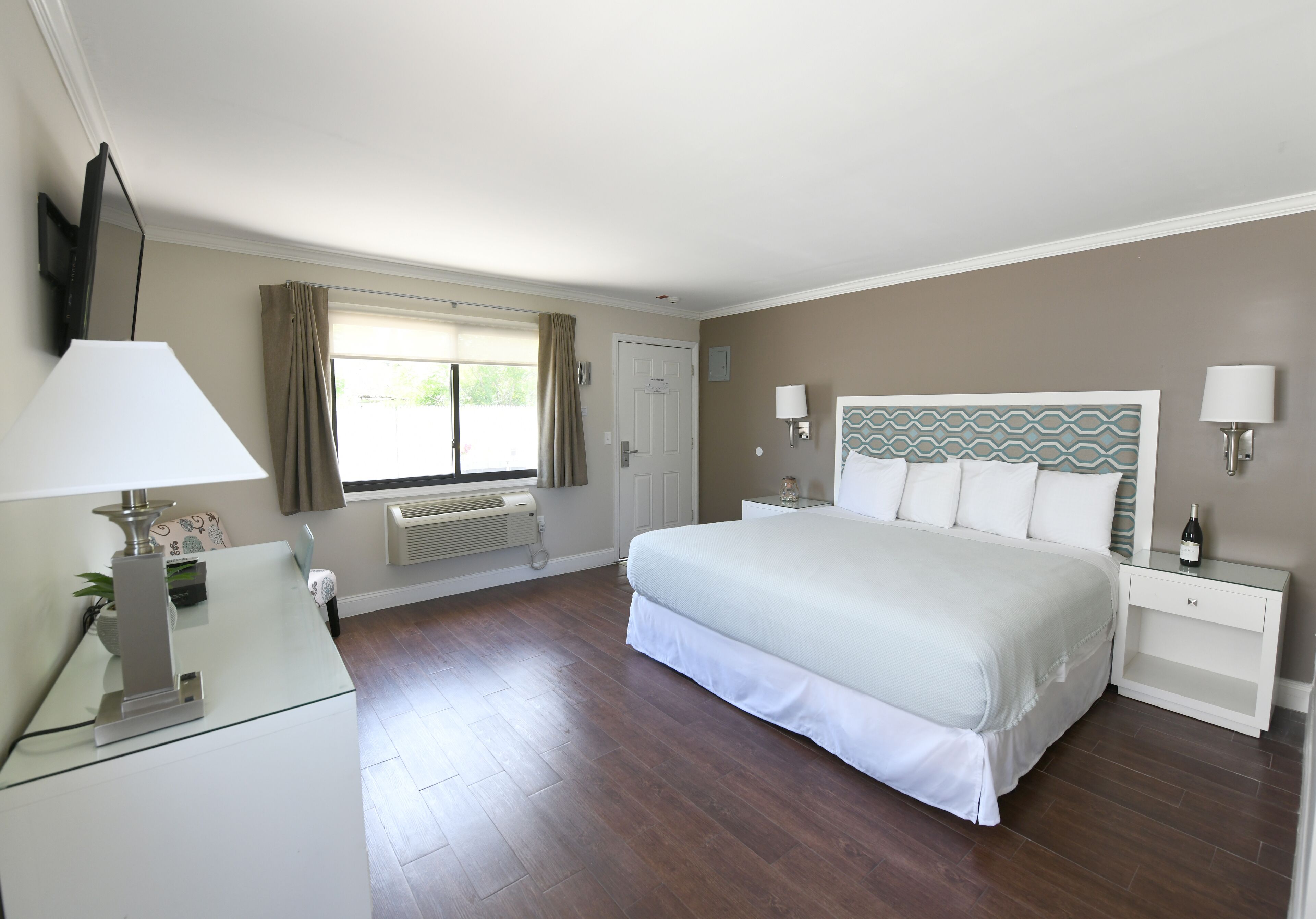 A bedroom with a large bed, a bedside table with a lamp, a nightstand, a window with curtains, and a wall-mounted TV.