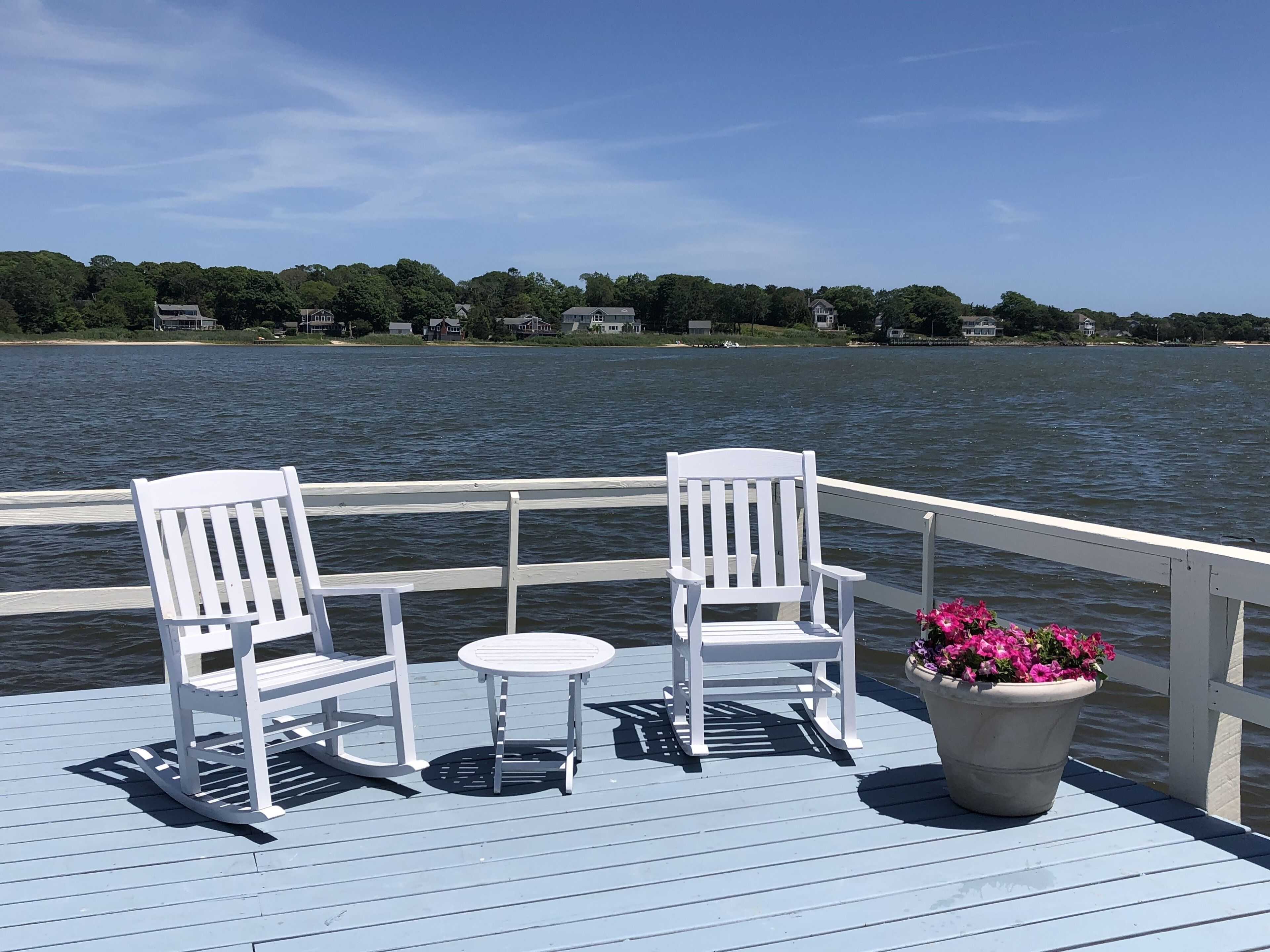 Una terraza de madera con dos sillas Adirondack blancas, una mesita redonda y una maceta con flores rosadas.
