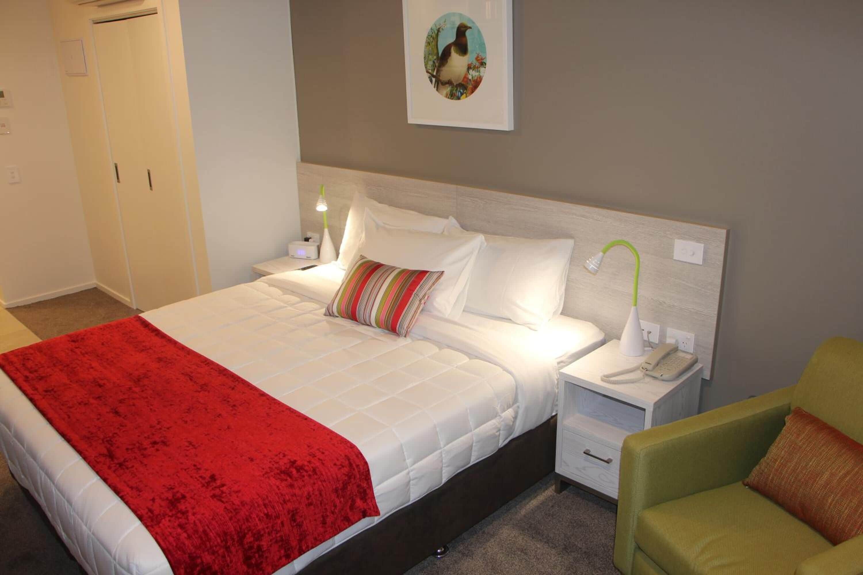 Habitación de hotel con cama, mesita de noche con lámpara, teléfono, una silla y un cuadro en la pared.