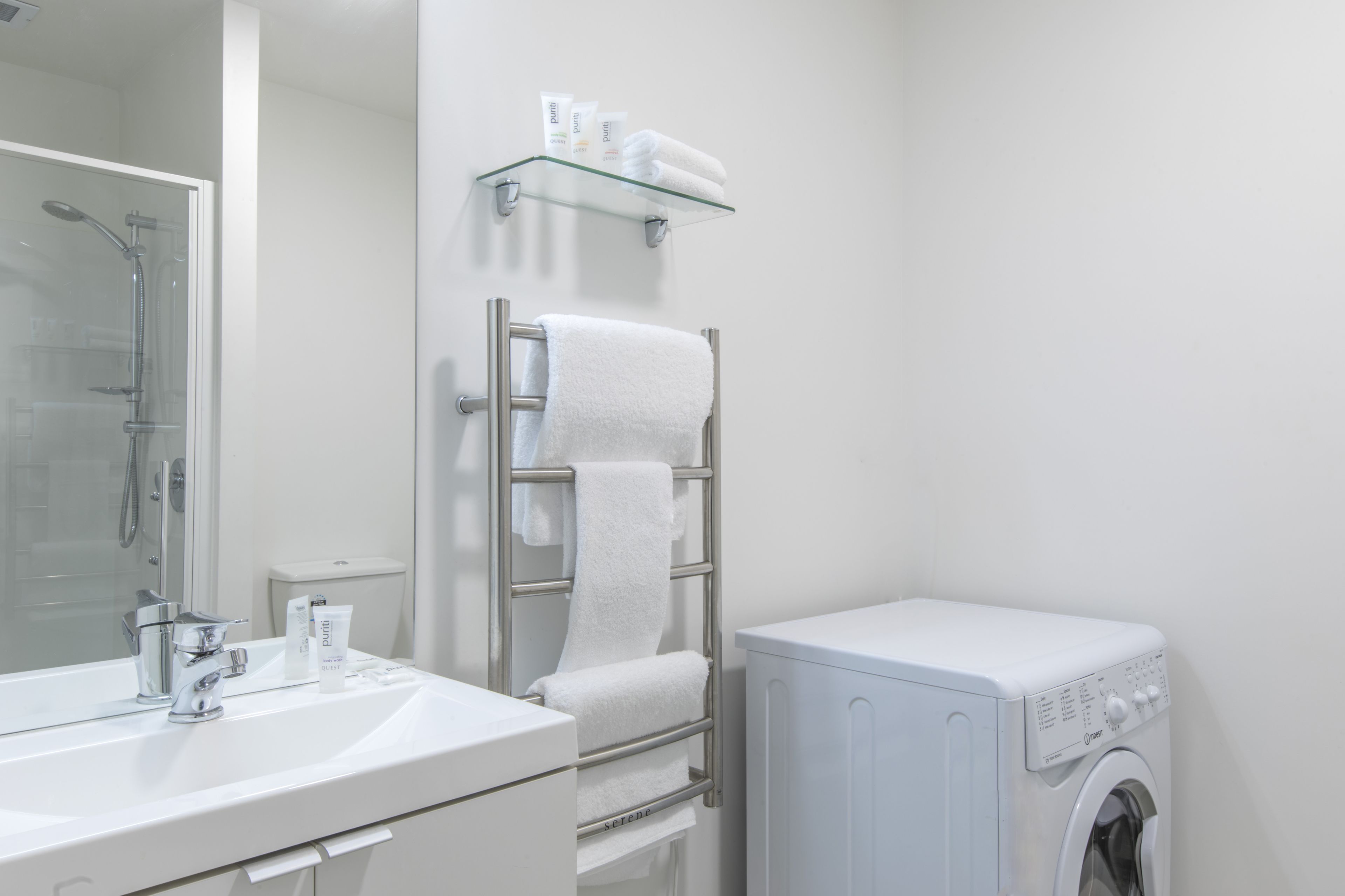 Un baño moderno con repisas de vidrio para artículos de aseo, un perchero con toallas, un lavamanos con espejo y una lavadora de carga frontal.