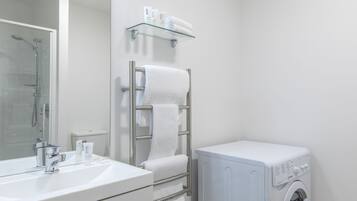 Un baño moderno con repisas de vidrio para artículos de aseo, un perchero con toallas, un lavamanos con espejo y una lavadora de carga frontal.