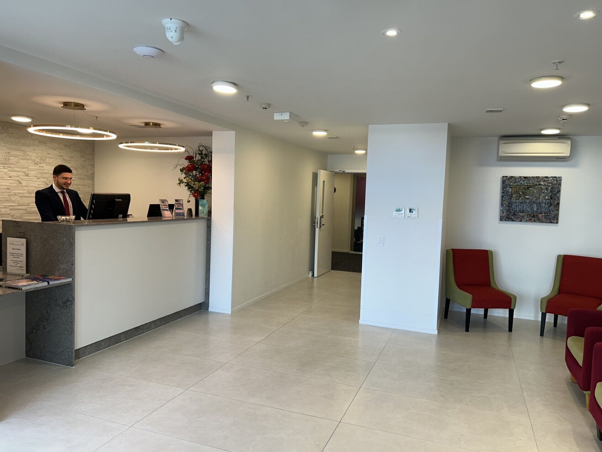 A hotel reception area with a reception desk, a man standing behind it, and two red chairs.