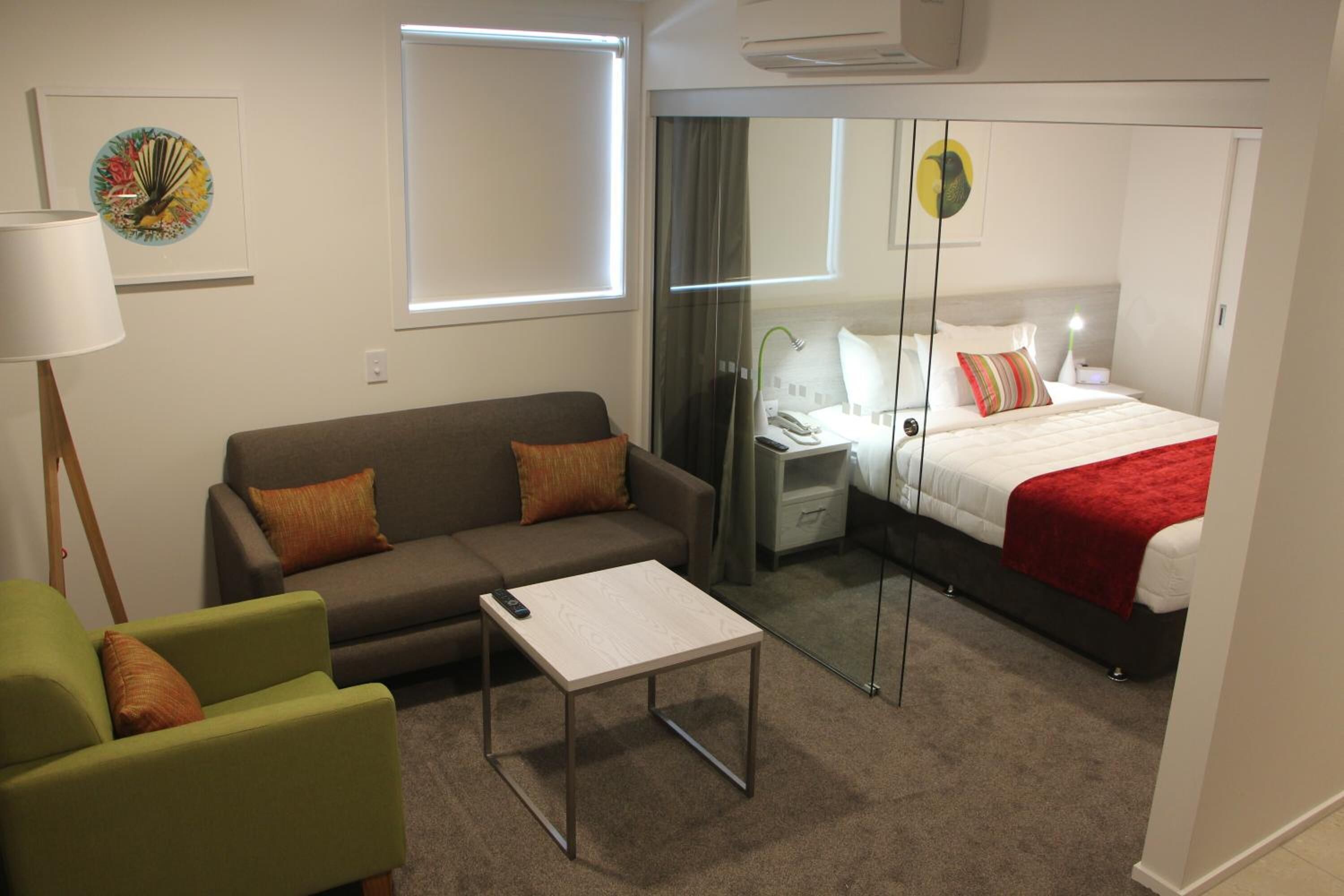 Une chambre d’hôtel moderne avec un canapé, une petite table, un lit recouvert d’une couverture rouge, une table de chevet avec une lampe et un miroir.