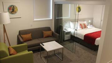 Une chambre d’hôtel moderne avec un canapé, une petite table, un lit recouvert d’une couverture rouge, une table de chevet avec une lampe et un miroir.