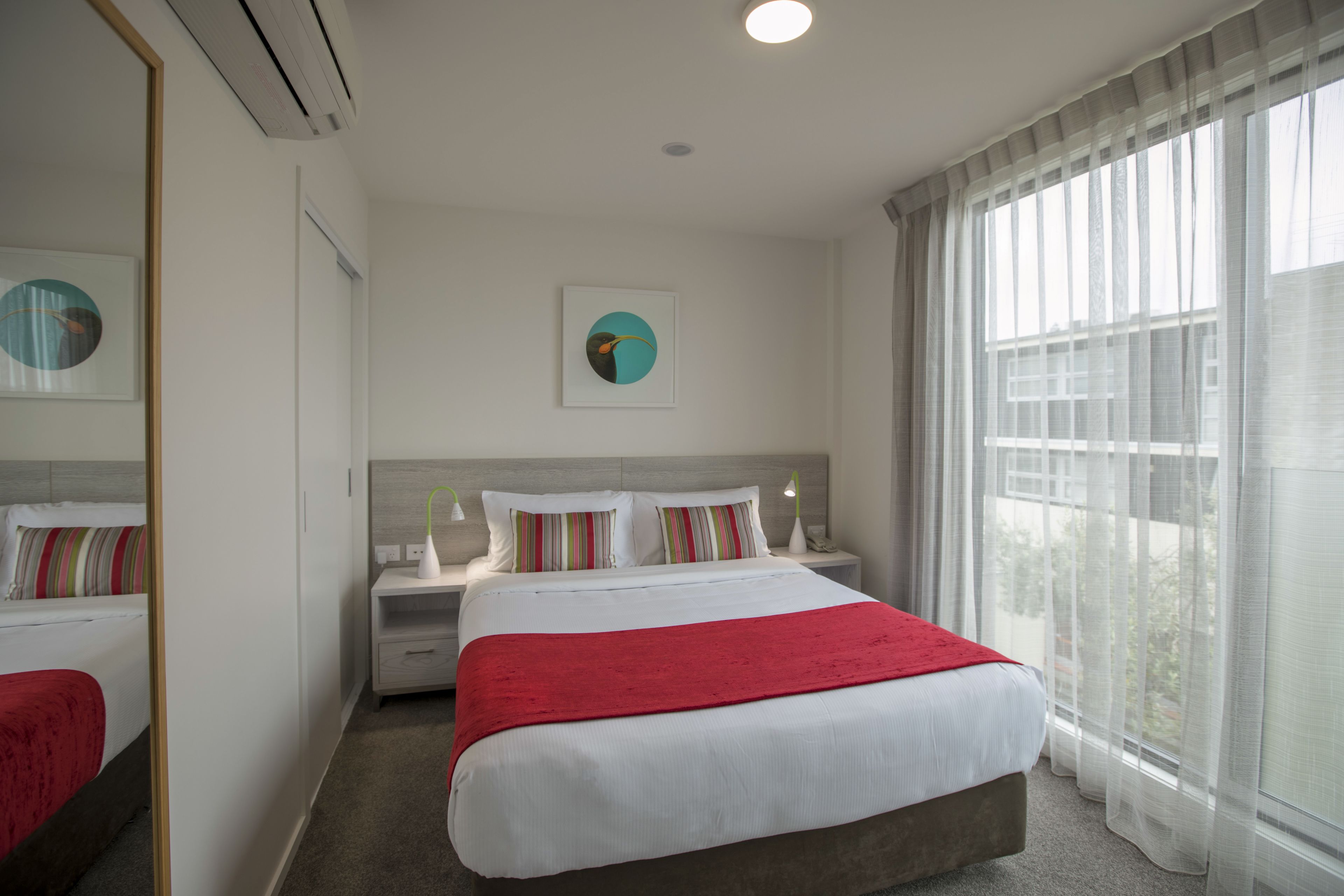 A hotel room with a large bed, a nightstand, a bedside table, a mirror, and a window with sheer curtains.