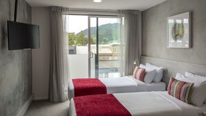 Une chambre d’hôtel avec deux lits, une télévision, une grande fenêtre avec des rideaux et une vue sur des immeubles et des collines.