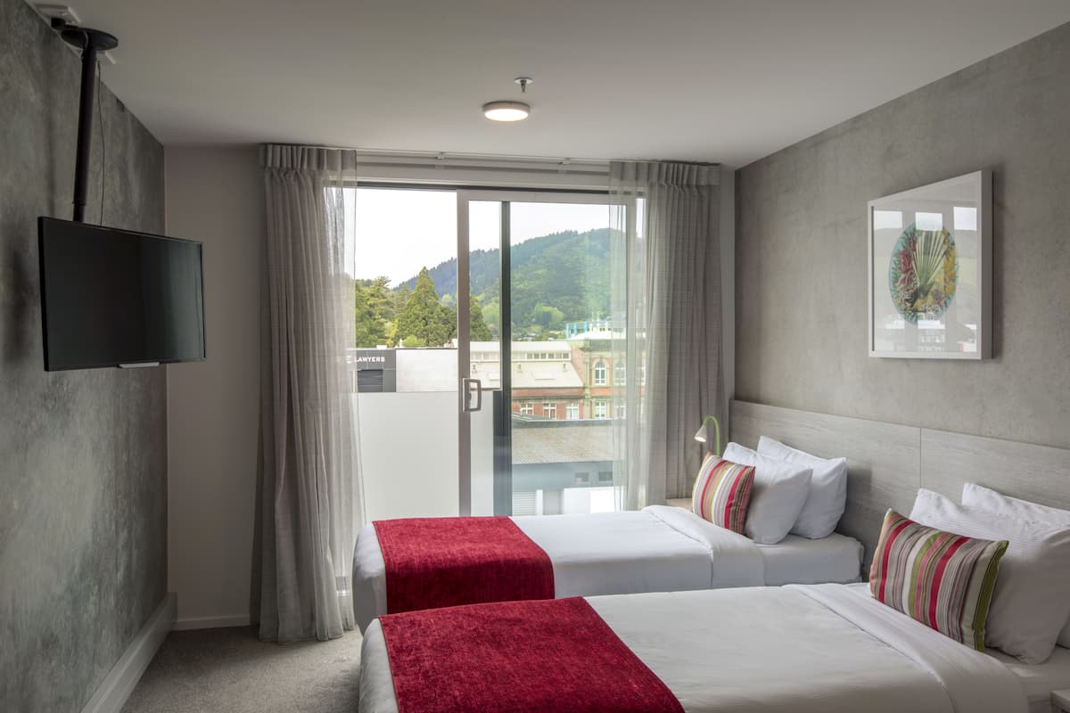 Habitación de hotel con dos camas, un televisor, un ventanal con cortinas y vistas a edificios y colinas.