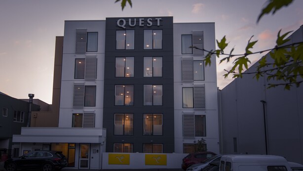 Un bâtiment sur lequel on peut lire l’inscription « QUEST » en haut, doté de nombreuses fenêtres et présentant un style architectural moderne.