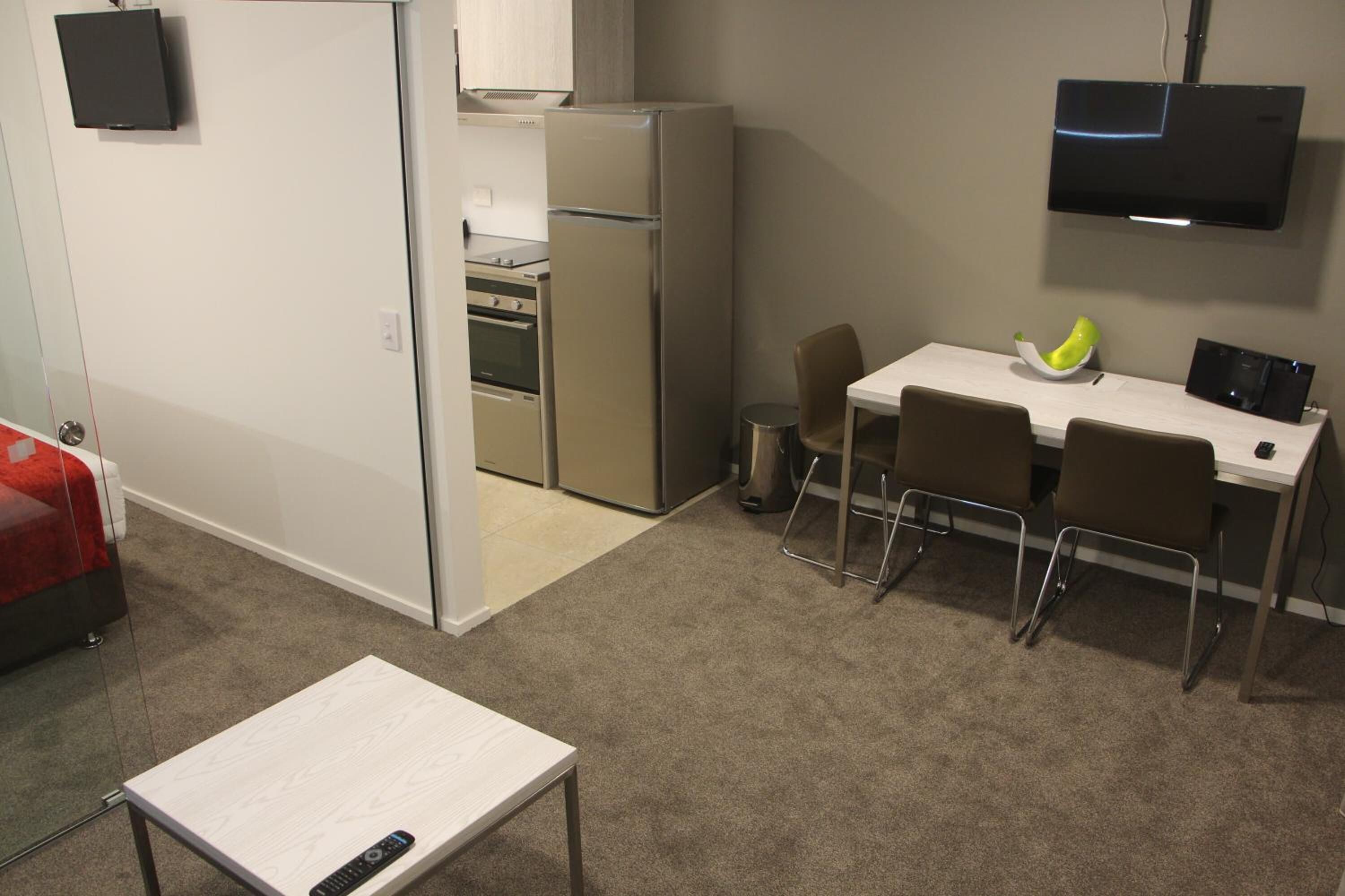 A compact kitchen with a refrigerator, stove, and microwave. There is a dining area with a table and chairs. A TV is mounted on the wall.
