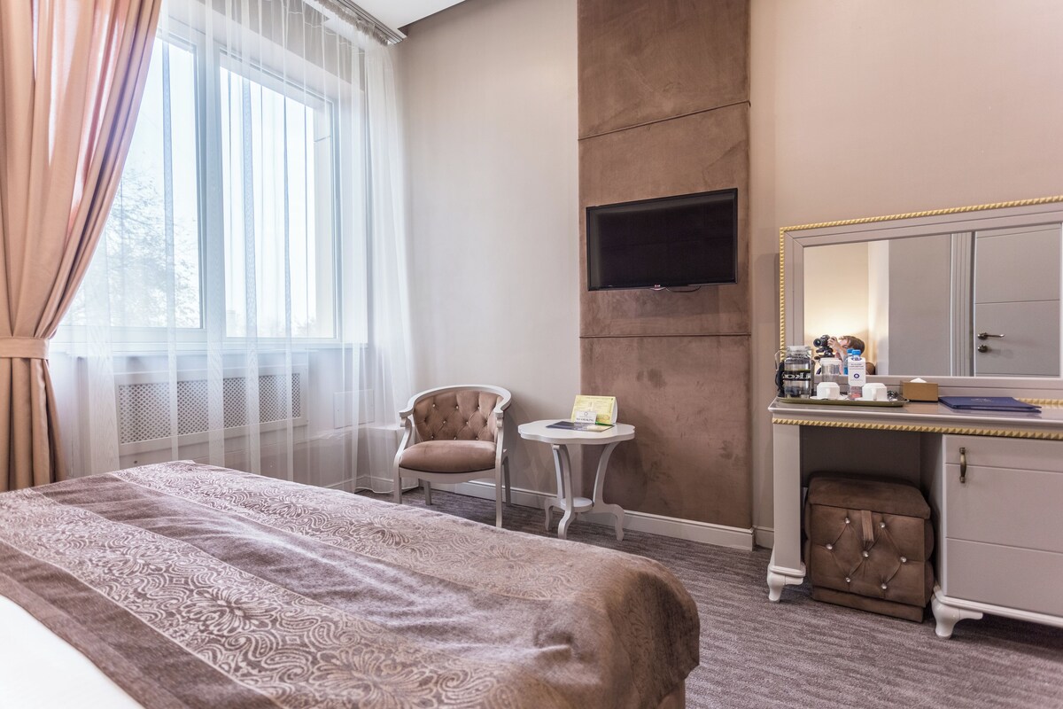 A hotel room with a bed, a chair, a small table, a TV, a mirror, and a dresser.